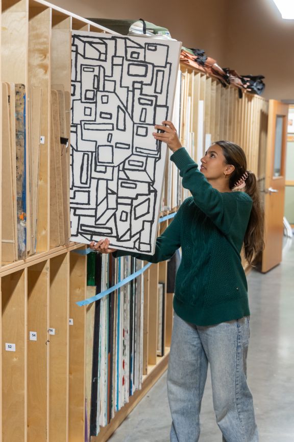 A girl pulling a large canvas from an art shelf