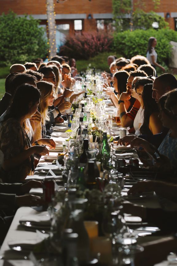 Many guests seated at an outdoor dining table in evening light chatting over drinks